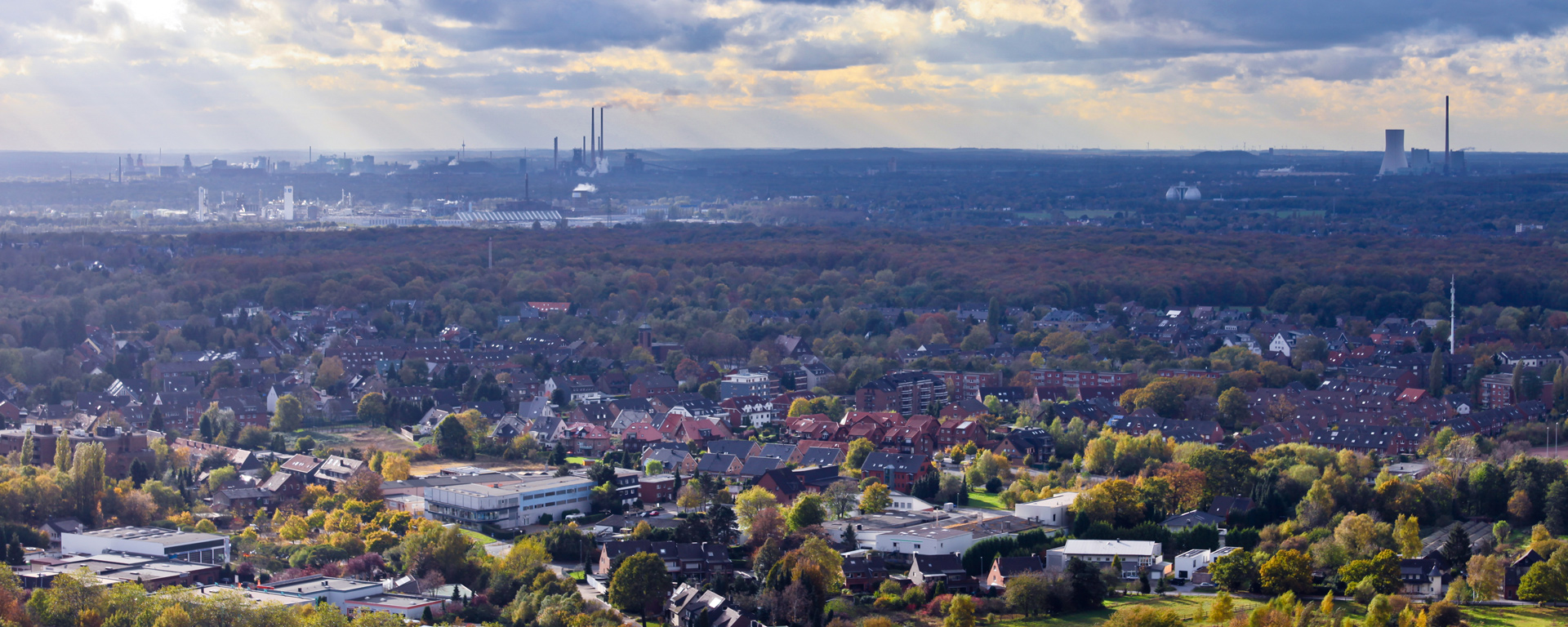 Luftaufnahme der Stadt Oberhausen mit Bäumen und Häusern vor aufragenden Industrieschloten und bewölktem Himmel